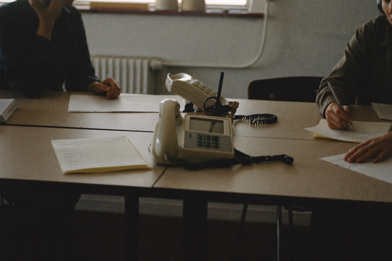 Two people at plain desks taking notes beside a landline telephone